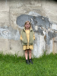 A child standing on grass against a wall, wearing a pistachio green wool vest over a yellow flower dress and blundstone shoes. The vest has a zipper and 2 pockets.