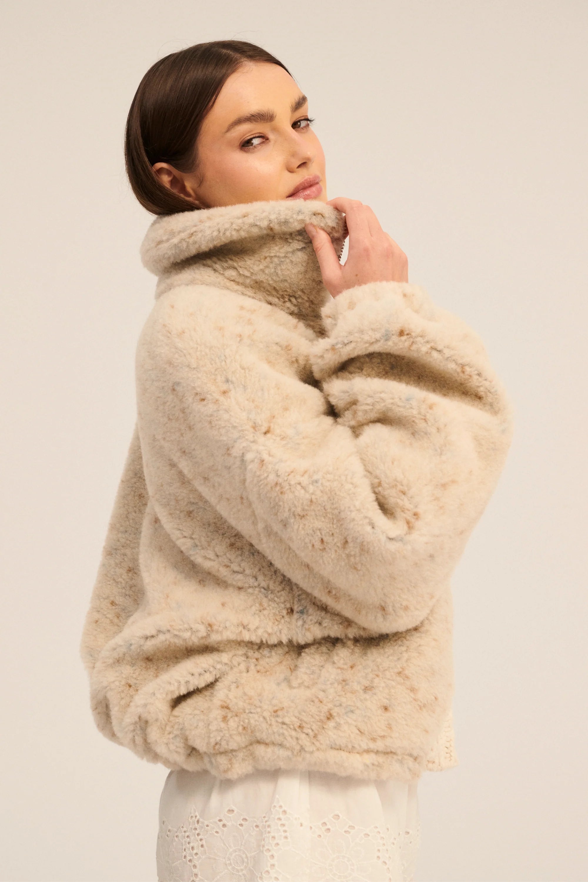 A woman wearing a beige, hip-length, sheepskin wool sweater with a triangle neckline and single-breasted velour buttons.