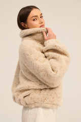 A woman wearing a beige, hip-length, sheepskin wool sweater with a triangle neckline and single-breasted velour buttons.